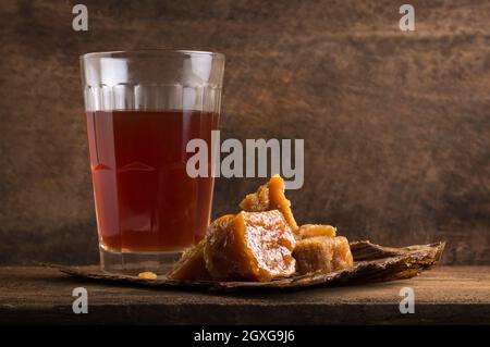 Stücke von gezacktem oder unraffiniertem Zucker oder Palmzucker mit einem Glas Tee, traditioneller Süßstoff in Südostasien mit einem heißen Getränk auf Holzoberfläche Stockfoto