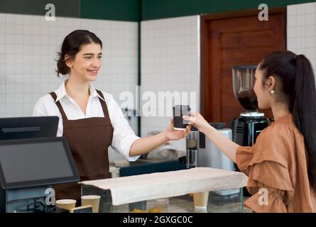 Der kaukasische Barista mit Schürze serviert jungen asiatischen Gästen heißen Kaffee. Morgendliche Atmosphäre in einem Café. Stockfoto