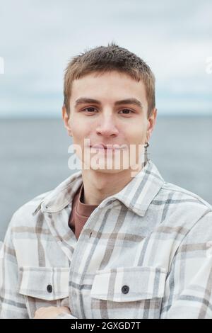 Vertikales Porträt eines lächelnden jungen Mannes, der die Kamera am Strand mit dem Himmel im Hintergrund betrachtet Stockfoto