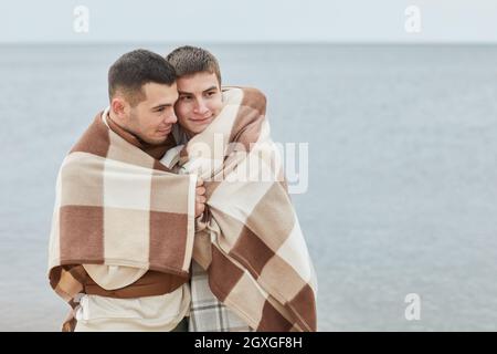 Minimalporträt eines jungen schwulen Paares, das sich am Strand umarmt, eingewickelt in eine Decke mit Wasser im Hintergrund, Kopierraum Stockfoto