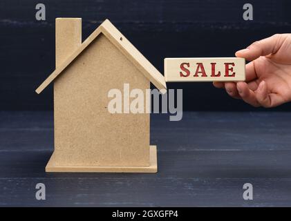 Holzmodell eines Hauses und eine Hand hält einen Block mit einer Inschrift Verkauf auf blauem Hintergrund. Hausverkauf Konzept, Immobilien-Investitionen, makler s Stockfoto