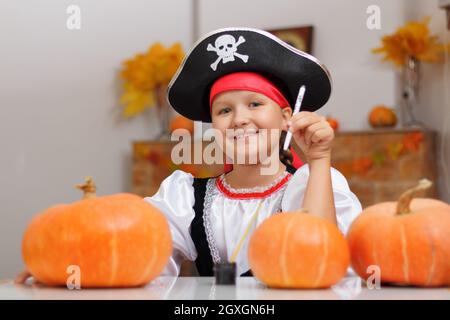 Halloween zu Hause feiern. Ein kleines Mädchen, das als Pirat gekleidet ist, sitzt am Tisch und zieht einen Kürbis an. Das Kind schaut in die Kamera. Stockfoto