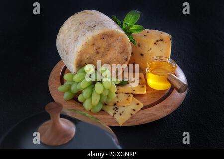 Hartkäse mit Trauben und Honig auf runder Holzplatte. Glasdeckel für Käseplatte. Foto auf schwarzem Hintergrund. Draufsicht. Stockfoto