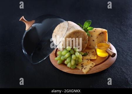 Hartkäse mit Trauben und Honig auf einem runden Holzteller. Glasdeckel für den Käseplatte. In Scheiben geschnittene Käsestücke. Foto auf schwarzem Hintergrund. Minze le Stockfoto