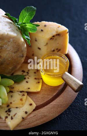 Hartkäse mit Trauben und Honig auf einem runden Holzteller. In Scheiben geschnittene Käsestücke. Foto auf schwarzem Hintergrund. Minzblätter. Draufsicht. Vertikales Foto. Stockfoto