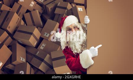 Der Weihnachtsmann zeigt einen leeren Platz für das Weihnachtsgeschenk an, mit fertigen Paketen im Hintergrund Stockfoto