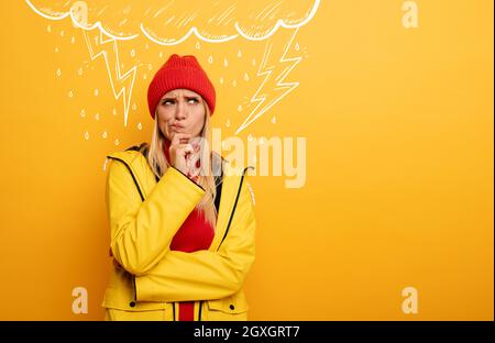 Blonde Girl denkt über negative Dinge nach. Wolke, Regen und Blitz über ihr. Verwirrter und nachdenklicher Ausdruck. Gelber Hintergrund Stockfoto