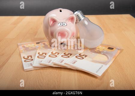 Konzept Energiepreis: Sparschwein mit Glühbirne und Geld auf Holzhintergrund. Stockfoto