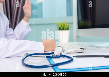 Mediziner arbeitet am Schreibtisch. Nahaufnahme des Stethoskops. Hand des Mannes Arzt Videoanruf Heben Sie die Hände, um Patienten auf Tisch Front Computer moni begrüßen Stockfoto