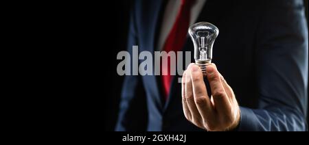 Geschäftsmann in schwarzem Anzug hält eine Glühbirne in der Hand auf einem schwarzen Hintergrund mit Kopierraum. Stockfoto