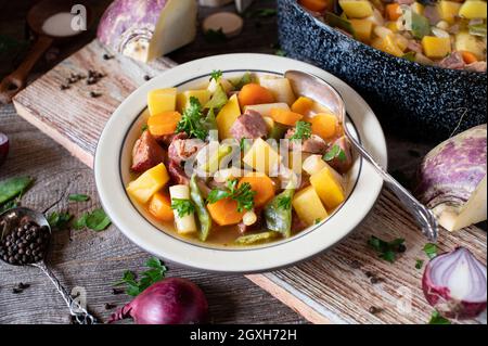 Herbstlicher Eintopf mit Rübe, Schwarzwurzel, Kartoffeln und Wurzelgemüse. Gekocht mit Schweinefleisch und serviert auf einem rustikalen Teller mit Topf auf Holztisch. Stockfoto