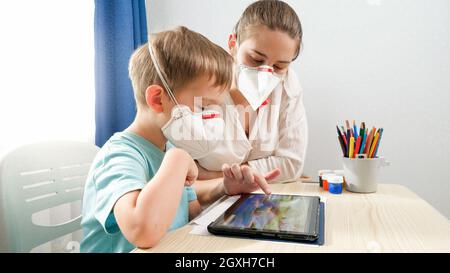 Mutter mit kleinen Sohn trägt schützende medizinische Atemschutzmasken mit Tablet-Computer. Hausaufgaben zu machen und zu Hause zu studieren während Lockdown und selbst Stockfoto