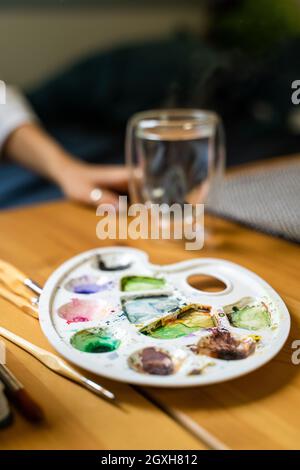 Mädchen in der Nähe von Aquarellen, Pinsel und Paletten auf dem Tisch neben warmem Wasser im Glas Stockfoto