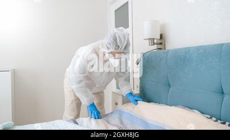 Junge Hausfrau in schützendem Biogefahrenanzug, die während der Covid-19-Pandemie und des Lockdowns Bett macht. Stockfoto