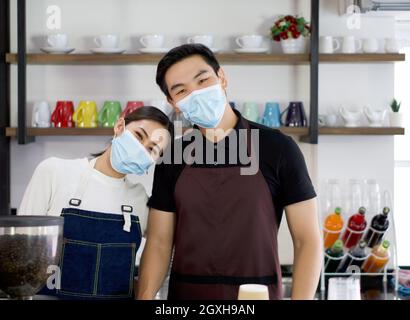 Der Ladenbesitzer und Barista, die eine Gesichtsmaske tragen, posiert fröhlich, bevor sie mit der täglichen Arbeit beginnen. Morgendliche Atmosphäre in einem Café. Stockfoto