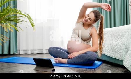 Lächelnde Schwangerin praktiziert Yoga online und schaut auf einen Tablet-Computer. Konzept eines gesunden Lebensstils, Gesundheitsfürsorge und Sport während der Schwangerschaft. Stockfoto