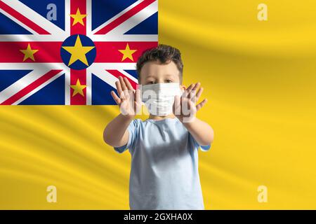 Kleiner weißer Junge in einer Schutzmaske auf dem Hintergrund der Flagge von Niue. Macht mit seinen Händen ein Stoppschild, bleibt zu Hause Niue. Stockfoto