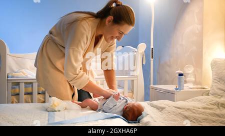 Lächelnde junge Mutter, die Windeln gewechselt und ihren neugeborenen Sohn abgewischt hat, wurde nachts auf dem Bett unordentlich. Schlaflose Nächte der Eltern und Babyhygiene. Stockfoto