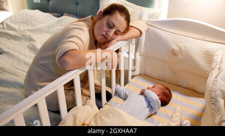 Die junge müde und erschöpfte Mutter schlief ein, während sie nachts die Krippe ihres neugeborenen Babys schaukelte. Konzept der schlaflosen Nächte und Elterndepression afte Stockfoto