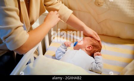 Fürsorgliche Mutter, die neben dem Kinderbett sitzt und ihr neugeborenes Kind streichelte, das im Kinderbett liegt. Konzept von glücklicher Elternschaft und Familienglück. Stockfoto