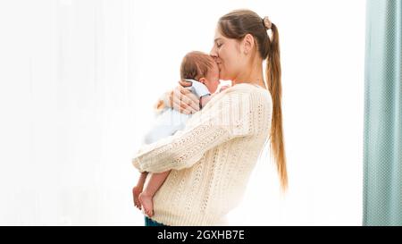 Glücklich lächelnde junge Mutter kuscheln und küssen ihren neugeborenen Jungen gegen helle Sonne scheint durch Fenster. Konzept von Familie Glück und Liebe Stockfoto