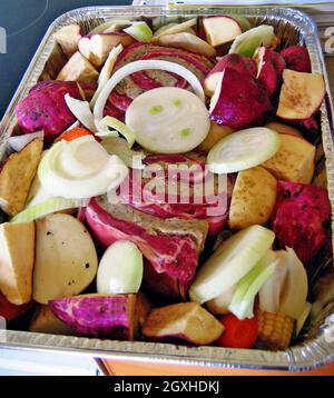 Ein Aluminiumtablett ist mit Speisen gefüllt, die für eine traditionelle Außenhangi zubereitet werden. Die Zwiebeln, das gerollte Flankensteak, die Kartoffeln und andere Lebensmittel werden mit Aluminiumfolie bedeckt und in einen dampfgespeisten Hangi gelegt. Stockfoto