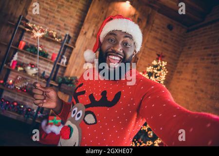 Foto von lustigen süßen dunklen Haut Mann tragen roten Pullover hält Feuerwerk Stick tacking Selfie lächelnd drinnen Zimmer Haus Stockfoto