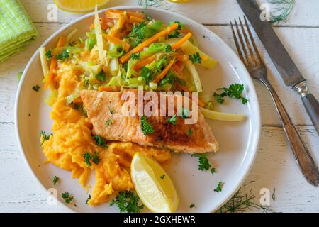 Gericht mit gebratenem Lachsfilet in der Pfanne und Süßkartoffelpüree, serviert mit gemischten Gemüsestreifen auf einem Teller auf einem weißen Holztisch. Ansicht von oben Stockfoto