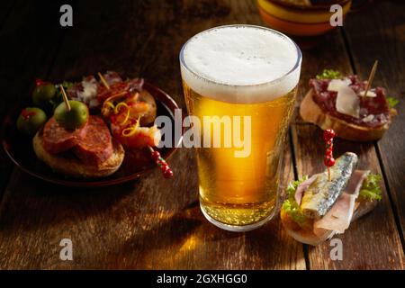 Leckere Auswahl an herzhaften spanischen Tapas, serviert mit einem kalten Pint Bier mit schaumigen Kopf auf einer rustikalen schattigen Holztheke in einer Taverne in einem hohen Angl Stockfoto