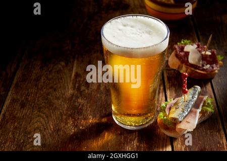 Ein Pint kaltes Bier vom Fass in einem hohen Glas mit leckeren Tapas auf frischem Baguette, serviert auf einer rustikalen Theke mit Copyspace in einem Pub oder einer Taverne Stockfoto