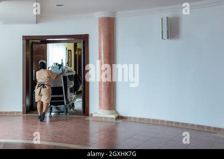 Das Zimmermädchen schob den Wagen durch den Flur im Hotel. Zimmermädchen bei der Arbeit und Trolley mit Zimmerbedarf und Reinigungsausrüstung im Hotel Stockfoto