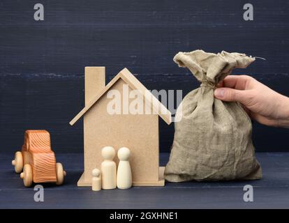 Die Hand hält einen Leinwandbeutel voller Geld und ein Holzhaus auf blauem Hintergrund. Das Konzept des Immobilienkaufs, der Vermietung. Rentable Investition i Stockfoto