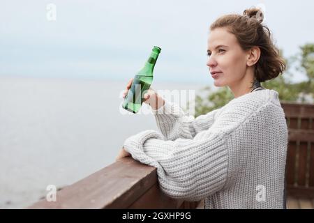 Minimale Seitenansicht Porträt einer jungen Frau, die beim Biertrinken am Strand den Blick aufs Meer genießt, Kopierraum Stockfoto