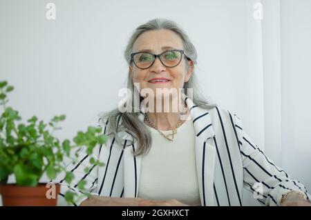 Ältere grauhaarige Geschäftsfrau in gestreifter Jacke mit Brille arbeitet in ihrem Büro am Schreibtisch sitzend. Glücklicher Ruhestand, Beschäftigung und Stockfoto