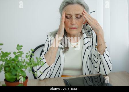 Müde ältere grauhaarige Geschäftsfrau in gestreifter Jacke mit Brille arbeitet in ihrem Büro am Schreibtisch sitzend und fühlt sich aufgrund der Wechseljahre schlecht Stockfoto