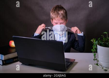 Ein Junge in einem Schulanzug mit einer medizinischen Maske auf seinem Gesicht an einem Computer, kopieren Raum auf einem dunklen Studiohintergrund Stockfoto