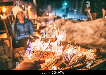 Winter Urlaub Skigebiet Frau rösten Marschallows in BBQ firepit Afterski Spaß Freizeitaktivitäten mit Freunden. Ein paar, die das Marschmallchen am Spieß anmachen Stockfoto