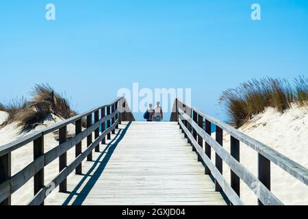 An einem Sommertag mit blauem Himmel laufen Touristen über einen Holzsteg zum Dünenstrand von Aveiro. Stockfoto