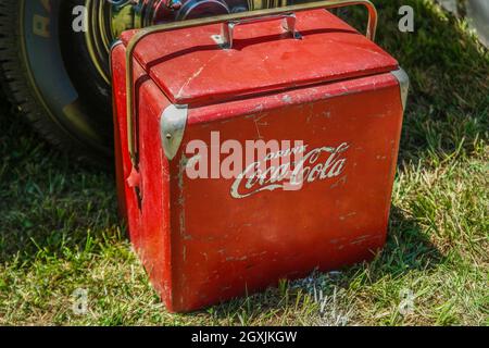 Ein antiker Coca-Cola Reisekühler aus Metall im Retro-Stil mit Patina, die auf dem Boden neben einem Auto im Freien sitzt, aus der Nähe der Sichtwinkel Stockfoto