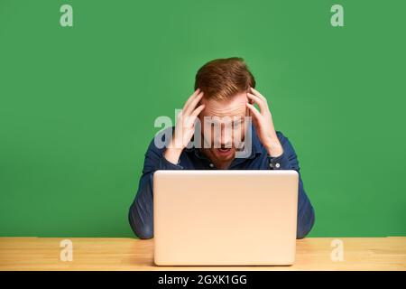 Schockiert jungen rothaarigen Geschäftsmann im Hemd halten Kopf in den Händen und lesen Internet-Nachrichten auf Laptop Stockfoto