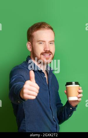 Porträt eines erfolgreichen Rotschopf-jungen kaukasischen Mannes mit Bart, der Kaffee zum Mitnehmen hält und den Daumen nach oben zeigt Stockfoto