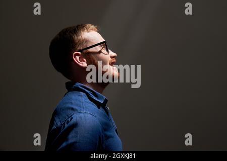 Seitenansicht eines lächelnden, hübschen jungen rotbärtigen Mannes in einer Brille, der in einem dunklen Raum Licht ansah, Ambitionen Konzept Stockfoto