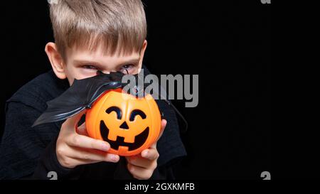 Ein Kind, das einen orangefarbenen Kürbis-förmigen Korb mit einem grinsenden Gesicht, Jacks Laterne und einer Fledermaus auf schwarzem Hintergrund hält. Der Junge wartet auf Halloween Stockfoto