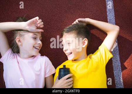 Fröhliche Sportkinder Bruder und Schwester Zwillinge liegen auf der Laufstrecke im Stadion, lächeln und schauen sich die Kamera von oben an. Teenager Mädchen und Jungen Athleten liegen auf Stockfoto