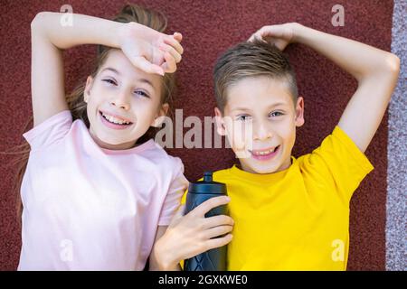 Kinder Sport Lifestyle-Thema. Fröhliche junge und junge Teenager liegen freudig auf der Gummiabdeckung der Laufstrecke des Stadions und löschen mit einem Lächeln in die Kamera Stockfoto