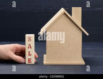Holzmodell eines Hauses und eine Hand hält einen Block mit einer Inschrift Verkauf auf blauem Hintergrund. Hausverkauf Konzept, Immobilien-Investitionen, makler s Stockfoto