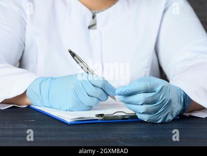 Ein Arzt in einem weißen Mantel und blauen medizinischen Handschuhen sitzt am Tisch und schreibt mit einem Stift aus nächster Nähe Stockfoto