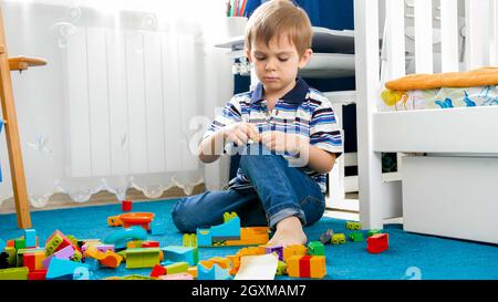 Porträt eines kleinen konzentrierten Jungen, der auf dem Teppich mit Spielzeugblöcken blassend ist. Stockfoto