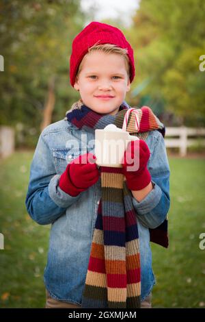 Hübscher junger Junge trägt Weihnachtskleidung, hält heißen Kakao mit Marshallows und Candy Cane draußen. Stockfoto