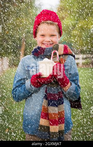 Hübscher junger Junge trägt Weihnachtskleidung und hält heißen Kakao mit Marshallows und Candy Cane draußen bei leichtem Schneefall. Stockfoto